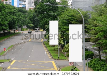 Hanging posters by the road in the city; blank vertical advertising banners on street lampposts, against lush green trees and plants. For OOH out of home template mock up. Royalty-Free Stock Photo #2279892591