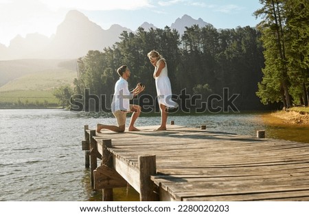 Man, woman and engagement proposal by lake on vacation with surprise, wow or happiness in sunshine. Couple, marriage offer and ring in nature for romance, love and happy on holiday in summer by water Royalty-Free Stock Photo #2280020203