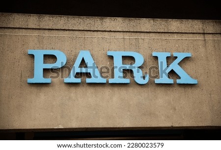 A green and white square sign with "PARK HERE" in bold black letters and a car graphic, mounted on a tall metal post against a backdrop of trees.