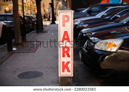 A green and white square sign with "PARK HERE" in bold black letters and a car graphic, mounted on a tall metal post against a backdrop of trees.