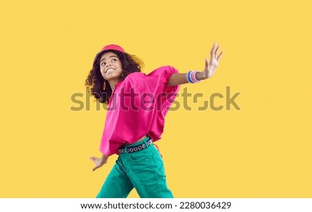 Happy child dancing. Cheerful little African American girl wearing loose oversized fuschia top and green jeans with trendy eyelet belt dancing isolated on solid yellow background. Kids fashion concept