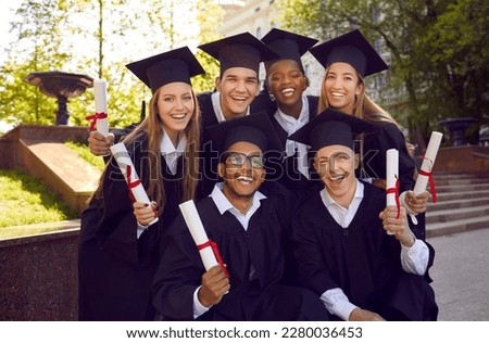 Obtaining higher education. Group portrait of happy excited graduates in academic gowns and caps holding diploma scrolls. Multiracial male and female university graduates pose together for photo.