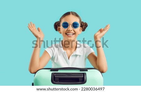 Overjoyed teenage girl with suitcase excited about summer vacation. Smiling teen kid in sunglasses isolated on blue studio background dream of summertime travel or holidays. Children and tourism. Royalty-Free Stock Photo #2280036497