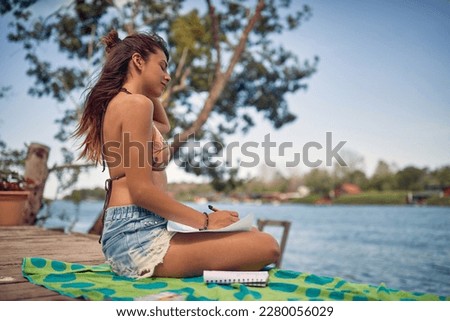 Young woman sitting on wooden jetty alone by water, holding pen looking at paper. Holiday, education, relaxation, lifestyle concept.