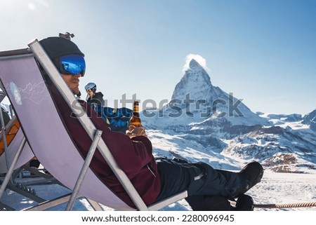 Young snowboarder cheering with a beer after skiing day in a bar or a cafe at the Zermatt ski resort in Swiss Alps with a fabulous view of the Matterhorn. Winter vacation concept.