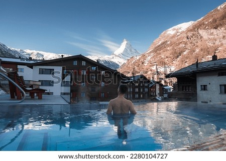 Young man swimming inside the luxury infinity outdoors pool with an amazing view at the Matterhorn peak in Zermatt, Switzerland. 