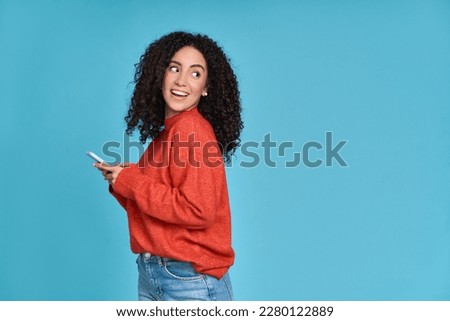 Young curious smiling happy pretty latin woman holding mobile phone, doing online shopping on cell, using apps on cellphone looking aside at copy space standing isolated on blue background. Royalty-Free Stock Photo #2280122889