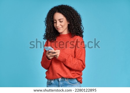 Young adult smiling happy pretty latin woman holding mobile phone looking at smartphone, typing message doing ecommerce shopping on cell, using trendy apps on cellphone isolated on blue background. Royalty-Free Stock Photo #2280122895