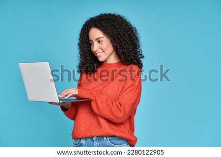 Young latin woman student using laptop device standing isolated on blue background. Smiling female model user holding computer, typing, surfing, searching job online or shopping website.