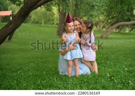 The mother hugs her daughters tightly and laughs loudly squeezing them with love. The youngest girl celebrates her third birthday outdoors in the park.