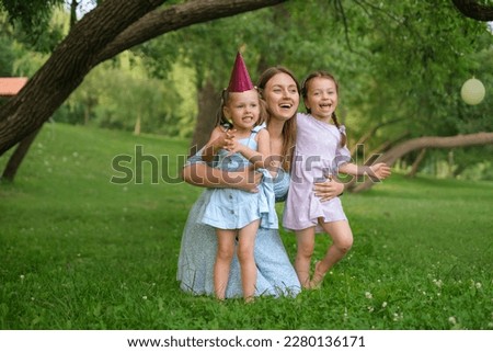Mom hugs her daughters and laughs merrily sitting in the park on the lawn. A birthday girl with a festive hat on her head. Mothers Day.