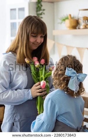 Cute little girl, baby daughter giving pink tulip flowers to her happy young beautiful mom. Celebration of Mother's or Women's day, birthday or anniversary. Cozy home atmopshere, kitchen. Springtime