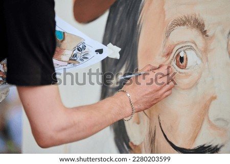 Young man artist draws with paint brush surreal man portrait on white canvas at art painting festival, paintings art picture process. Woman paints Salvador Dali portrait, art of atmospheric surrealism