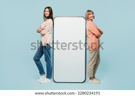 Full body sideways fun elder parent mom with young adult daughter two women together in casual clothes big huge blank screen area mobile cell phone isolated on plain blue background Family day concept
