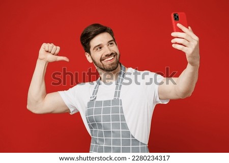 Young male housewife housekeeper chef cook baker man wear grey apron do selfie shot on mobile cell phone post photo point thumb finger on himslef isolated on plain red background. Cooking food concept