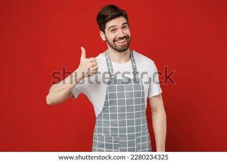 Young smiling happy fun cool satisfied male housewife housekeeper chef cook baker man in grey apron showing thumb up like gesture isolated on plain red color background studio. Cooking food concept