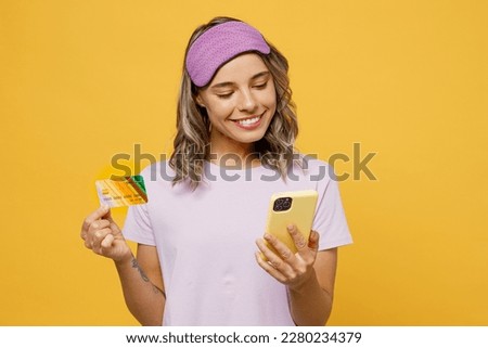 Calm young woman wear purple pyjamas jam sleep eye mask rest relax at home using mobile cell phone hold credit bank card order delivery isolated on plain yellow background Good mood night nap concept