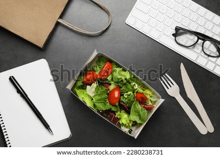 Container of tasty food, keyboard, cutlery and notebook on grey table, flat lay. Business lunch