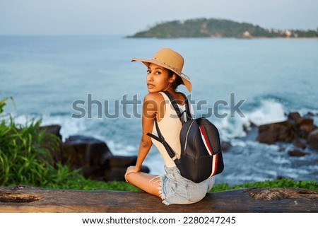 Multiethnic woman in straw hat enjoys tropical vacation sitting on bench at cliff with ocean view. Black female with backpack sightseeing on scenic location. Girl tanning on island on sea sunrise. Royalty-Free Stock Photo #2280247245
