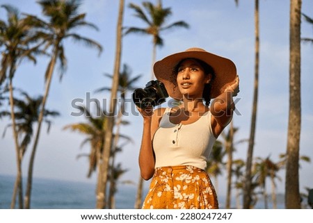 Young african female model tourist with camera in colorful clothes and straw hat taking pictures at tropical location at sunrise. Black travelling woman takes photos in exotic ocean scenery at dawn.