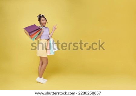 Cheerful happy thai asian woman enjoying shopping, she is carrying shopping bags pointing finger isolated on copy space yellow background.