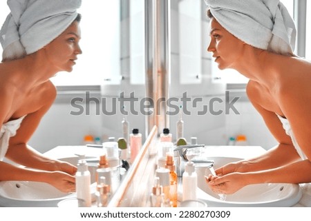 A beautiful woman in a white towel standing at the washbasin and mirror washing herself with tap water. Morning skin care routine. Cosmetology and spa procedures.