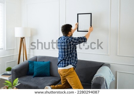 Rear view of a young man putting up a picture on the wall and decorations in his new living room home after moving