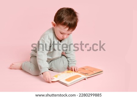 Happy toddler baby is reading a book on studio pink background. Child boy plays with a book. Kid age one year eight months, full height