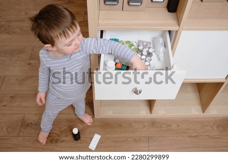 Toddler baby opened the cabinet drawer with pills and medicine. Child boy holding a pack of pills in the home living room. Kid age one year nine months Royalty-Free Stock Photo #2280299899
