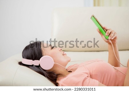 Peaceful girl in modern wireless headphones sit relax on comfortable couch listening to music looking at her smartphone