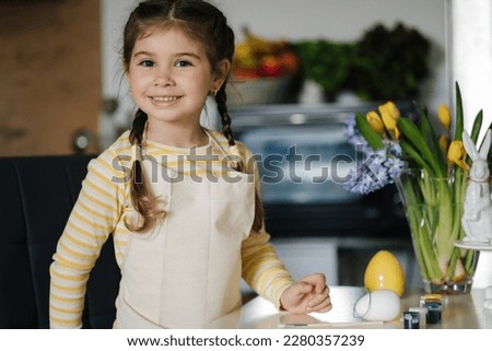 Adorable smiled girl hold brush and eggs in hands and preparing for painting. Easter decoration on kitchen