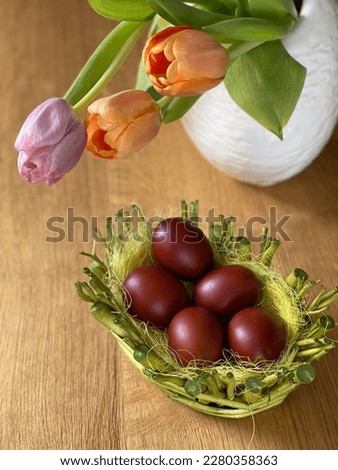 Close-up, almost top down of brown coloured Easter eggs in light green twigs basket and orange, pink tulips flowers in white vase, on oak wood. Eggs coloured by cooking with onion skins. 