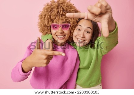 Portrait of cheerful young women friends make frame gesture with fingers capture best moment try to find perfect angle picture something dressed in casual jumper isolated over pink background.