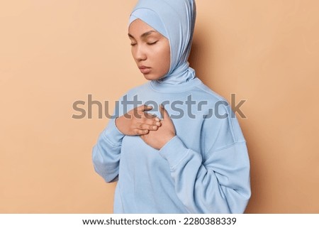 Calm thankful Muslim woman presses palms to chest keeps eyes closed expresses gratitude to you wears blue pullover and hijab wrapped on head isolated over brown background. Body language concept