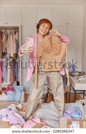 Happy redhead woman sells clothes during online stream holds mobile phone and fashionable jumper on hanger suggests to buy it poses on bed sorts out wardrobe stands against bedroom interior.