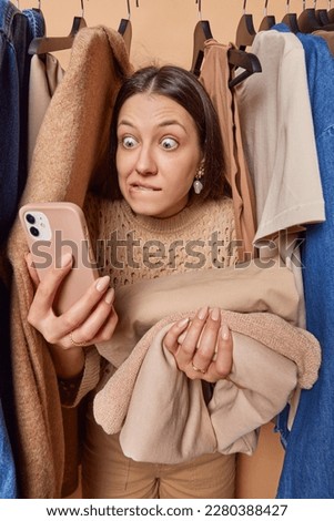 Surprised brunette woman stares at smartphone screen bites lips feels nervous holds folded clothes stands near clothes on hangers in wardrobe tries on clothes. Shopaholic and consumerism concept