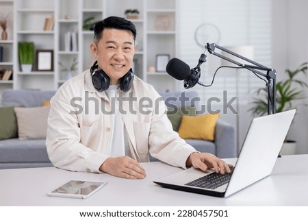 Portrait of blogger at home, Asian man smiling and looking at camera, man sitting at table with laptop in living room, recording audio podcasts and doing online radio.