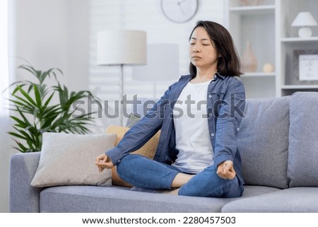 Young beautiful Asian woman alone at home, meditating sitting on sofa in living room in lotus position, sitting with eyes closed at home.