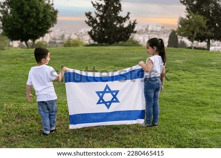 Young children stand on a hill holding an Israeli flag. A rear view little girl and boy with an Israel flag in the sunset. Royalty-Free Stock Photo #2280465415
