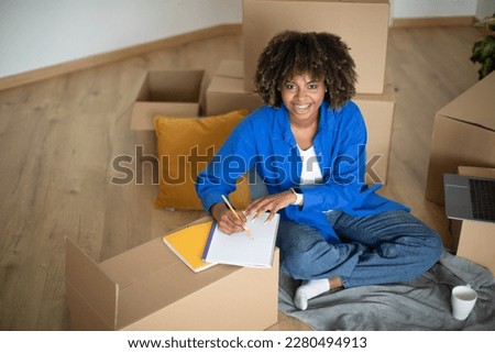 Shopping Planning. Happy Black Woman Making Checklist After Moving New Home, Above Shot Of Smiling Young African American Lady Sitting Among Cardboard Boxes With Belongings And Writing In Notepad