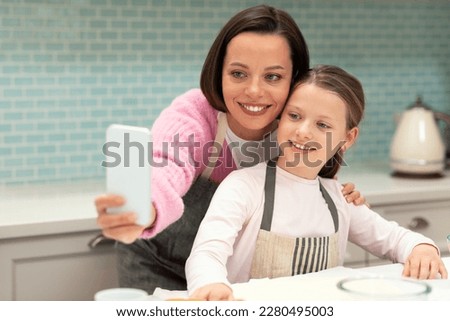 Smiling caucasian young mother and little daughter in aprons take selfie on phone, make cookies in kitchen interior. App for food blog at homemade sweets at home, shooting video, have video call Royalty-Free Stock Photo #2280495003