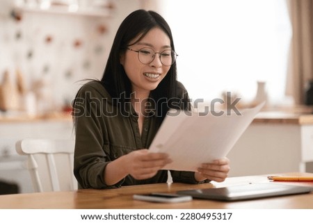 Happy young asian woman wearing eyeglasses sitting at kitchen desk with laptop and phone on, holding papers, checking correspondence or working with documents from home, copy space Royalty-Free Stock Photo #2280495317