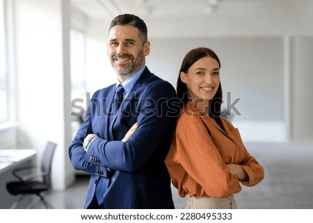 Two successful businesspeople standing back to back with arms crossed and smiling at camera, posing in modern office interior, copy space. Successful businessman and businesswoman Royalty-Free Stock Photo #2280495333