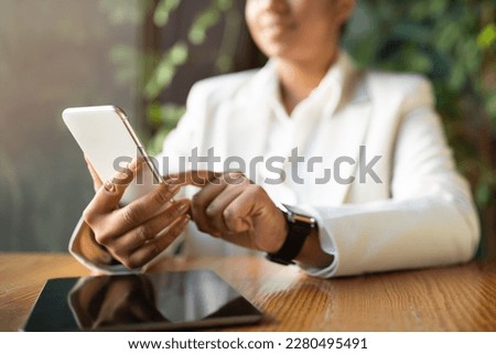 Happy young african american female in suit typing on phone with tablet with blank screen in modern eco cafe with green plants interior, cropped, blurred. App for work, study and business remotely