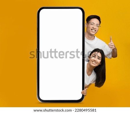 Online Offer. Smiling Asian Couple Peeking Out Behind Big Blank Smartphone And Showing Thumb Up Over Yellow Background, Happy Young Man And Woman Recommending New Mobile App Or Website, Mockup Royalty-Free Stock Photo #2280495581