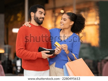 Husband Giving Credit Card For Shopping To Happy Wife Posing With Shopper Bags Standing At Modern Mall Outdoors. Multicultural Couple Spending Money On Weekend. Family Budget And Shopaholism