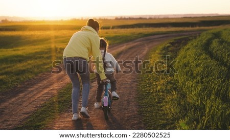 mom teaches ittle child ride bike summer road park sunset. happy family concept. cheerful child pedals bicycle sun. kid controls steering wheel two-wheeled bicycle summer sunset. weekends motherhood.