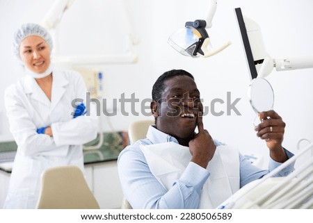 African-american man looking at his teeth through mirror after dental restoration procedure. Asian woman dentist standing in background and smiling.