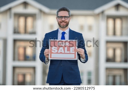 Successful real estate agent in a suit holding for sale sign near new apartment. Real estate agent with home loan contract, selling home. Realtor or real estate agent shows board for sale.