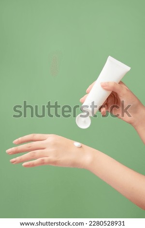A white tube without label is held in woman model’s hand with a cream texture on another hand. Green pastel background. Branding mockup Royalty-Free Stock Photo #2280528931